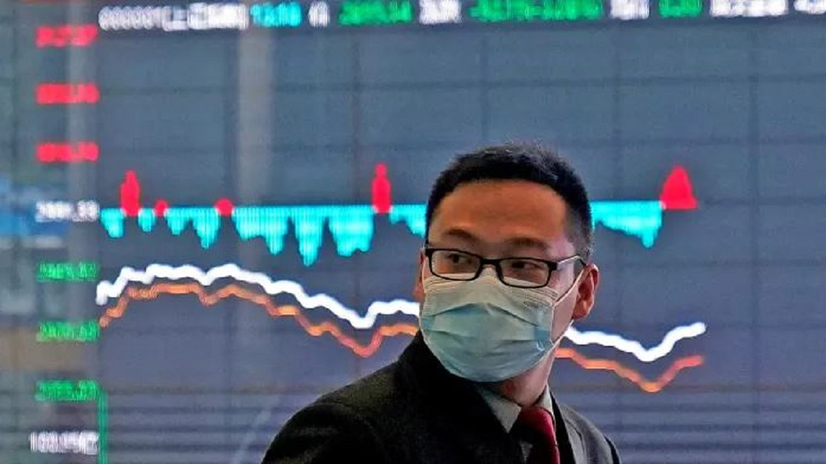 Man stands in front of stock market screens with mask on.