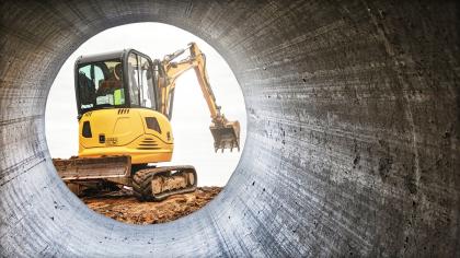 An excavator moves soil at a construction site framed by a large concrete pipe. 